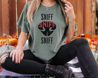 May include: A green t-shirt with the words "Sniff Sniff Sniff" in black and red lettering. The shirt is being worn by a person sitting on a patterned rug.