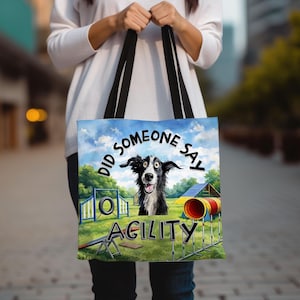 May include: A tote bag with a black and white dog design and the words "DID SOMEONE SAY AGILITY". The bag, held by a person, has black handles and depicts an agility course under a blue sky.