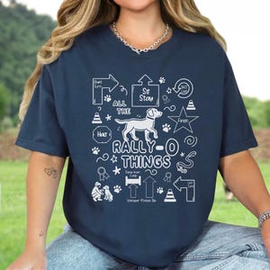May include: Navy blue t-shirt with a white graphic design featuring a dog, paw prints, and the text "ALL THE RALLY-O THINGS." The design includes arrows, stars, and other elements related to dog agility training. The shirt is a comfortable, casual style.