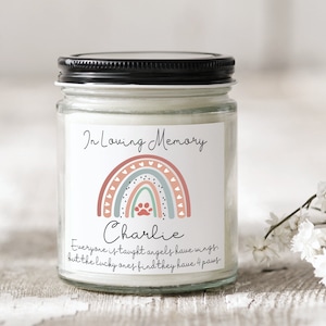 May include: A glass jar candle with a black lid and a white label. The label has a rainbow with hearts and a paw print in the center. The text on the label reads "In Loving Memory Charlie Everyone is taught angels have wings but the lucky ones find they have 4 paws".