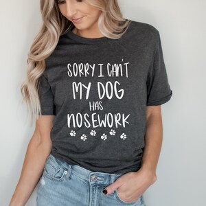 May include: Dark gray t-shirt with the text "SORRY I CAN'T MY DOG HAS NOSEWORK" in white, along with paw print graphics. The shirt is a casual style, suitable for dog lovers.