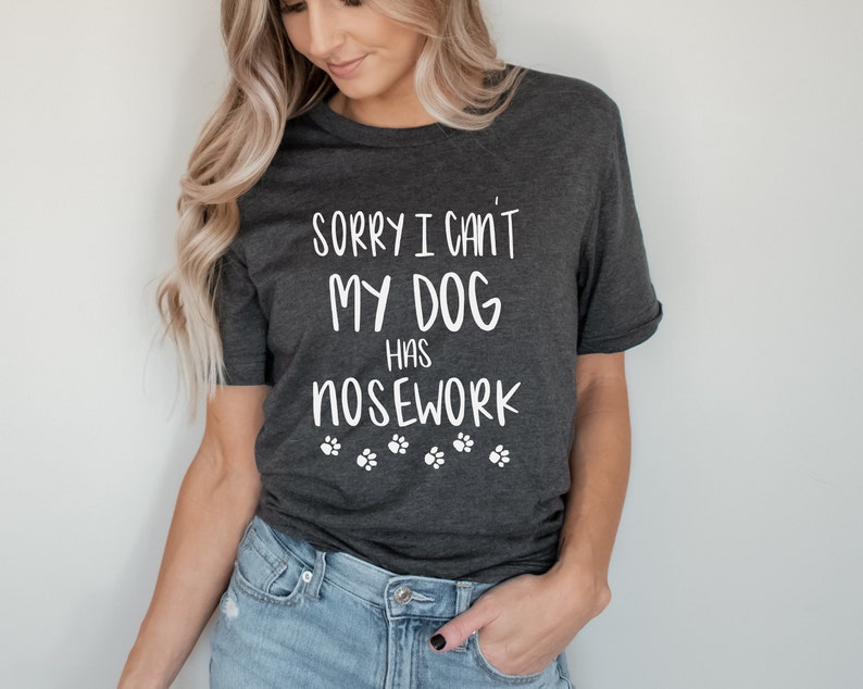 May include: Dark gray t-shirt with the text "SORRY I CAN'T MY DOG HAS NOSEWORK" in white, along with paw print graphics. The shirt is a casual style, suitable for dog lovers.