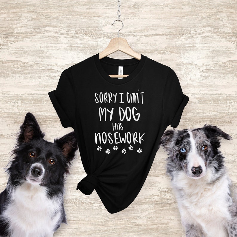 May include: Black t-shirt with the text "SORRY I CAN'T MY DOG HAS NOSEWORK" in white. The shirt is hanging on a wooden hanger. Two dogs, one black and white and the other gray and white, are on either side of the shirt.