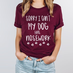 May include: A maroon t-shirt with the text "SORRY I CAN'T MY DOG HAS NOSEWORK" in white, along with paw print graphics. The shirt has rolled-up sleeves and is made of a soft material.