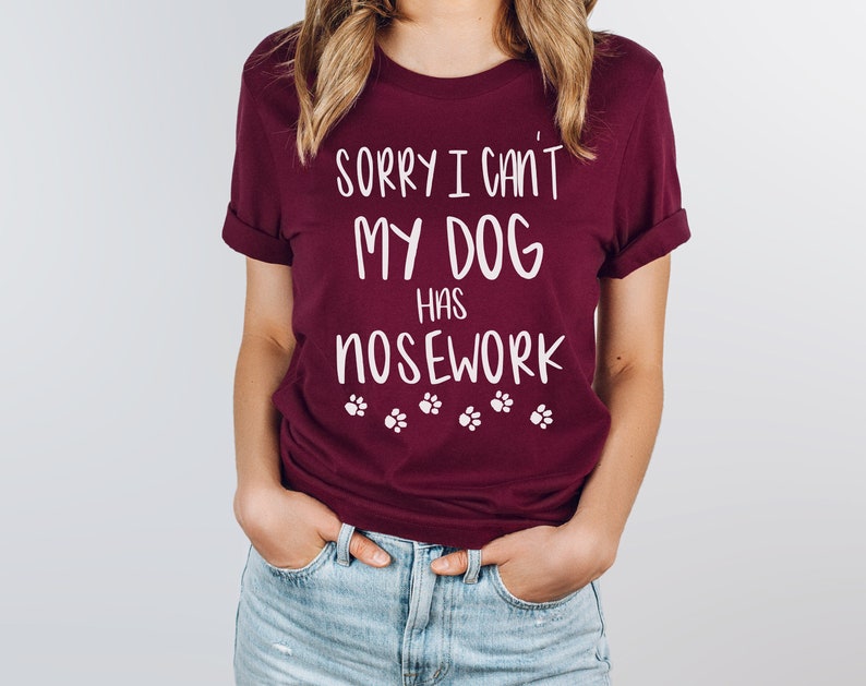 May include: A maroon t-shirt with the text "SORRY I CAN'T MY DOG HAS NOSEWORK" in white, along with paw print graphics. The shirt has rolled-up sleeves and is made of a soft material.