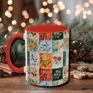 May include: A ceramic mug with a red interior and handle, featuring a patchwork design of embroidered flowers in various colors. The mug is white on the exterior with a floral quilt pattern. The background includes blurred lights and greenery.