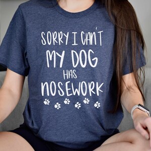 May include: A navy blue t-shirt with the text "SORRY I CAN'T MY DOG HAS NOSEWORK" in white, along with paw print graphics. The shirt is a crew neck style.