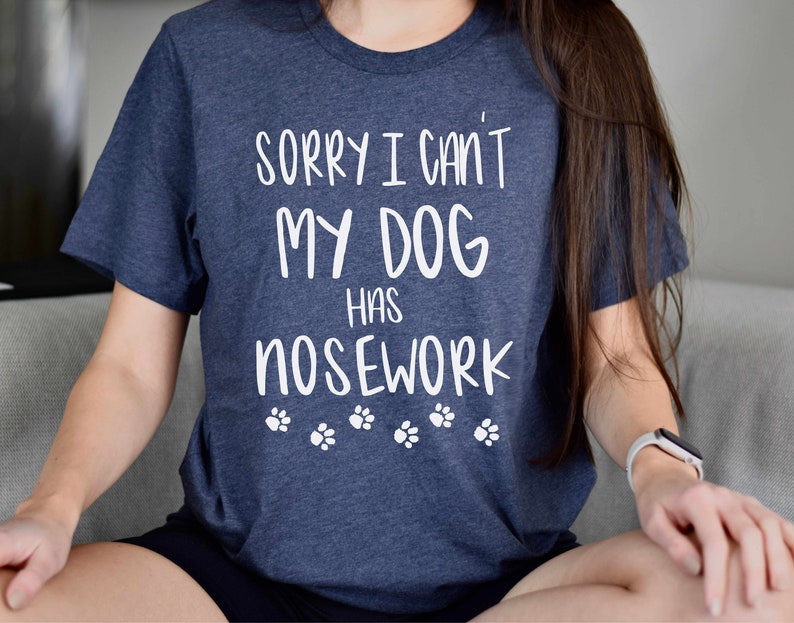 May include: A navy blue t-shirt with the text "SORRY I CAN'T MY DOG HAS NOSEWORK" in white, along with paw print graphics. The shirt is a crew neck style.