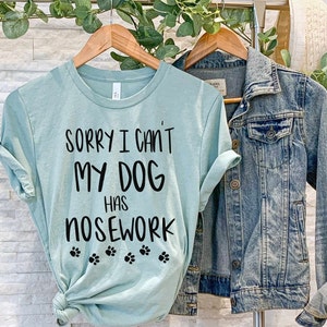 May include: A light teal t-shirt with the text "SORRY I CAN'T MY DOG HAS NOSEWORK" in black, along with paw prints. A denim jacket hangs next to the t-shirt. The t-shirt is on a wooden hanger.