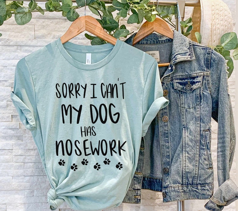 May include: A light teal t-shirt with the text "SORRY I CAN'T MY DOG HAS NOSEWORK" in black, along with paw prints. A denim jacket hangs next to the t-shirt. The t-shirt is on a wooden hanger.