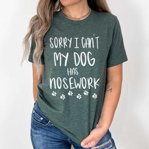 May include: Dark green t-shirt with the text "SORRY I CAN'T MY DOG HAS NOSEWORK" in white, along with white paw prints. The shirt is made of a soft material.