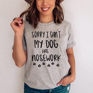 May include: A light gray t-shirt with the text "SORRY I CAN'T MY DOG HAS NOSEWORK" in black, along with paw print graphics. The shirt has rolled-up sleeves.