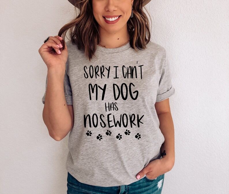 May include: A light gray t-shirt with the text "SORRY I CAN'T MY DOG HAS NOSEWORK" in black, along with paw print graphics. The shirt has rolled-up sleeves.