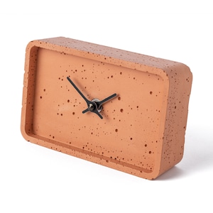 Op de afbeelding: Een rechthoekige terracotta-kleurige klok met zwarte wijzers. De wijzerplaat van de klok heeft een gespikkelde textuur.