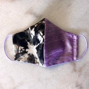 May include: A face mask featuring a split design with one side in a black and white abstract ink wash pattern and the other side in a solid textured purple fabric. The mask includes light purple elastic ear loops on both sides for secure attachment.