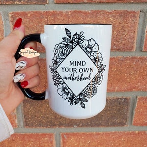 May include: White ceramic mug with a black handle and black rim. The mug has a black and white floral design with a diamond shape in the center that says "Mind your own motherhood".