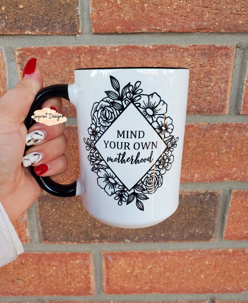 May include: White ceramic mug with a black handle and black rim. The mug has a black and white floral design with a diamond shape in the center that says "Mind your own motherhood".