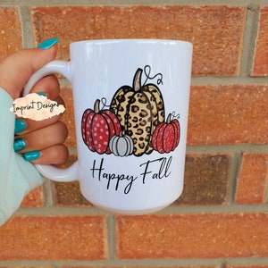 May include: White ceramic mug featuring a design of three pumpkins. The pumpkins are red, white and brown with a leopard print pattern. The text "Happy Fall" is printed below the pumpkins.