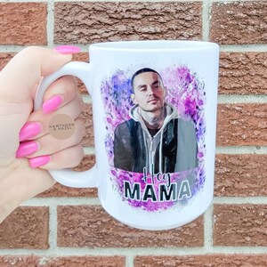May include: White ceramic mug featuring a watercolour-style image of a man with a tattoo on his neck. The image is surrounded by purple and pink splatters. The text "Hey Mama" is printed in black below the image.