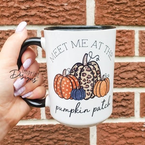 May include: White ceramic mug with a black rim and handle. The mug features a graphic of pumpkins with orange, brown, and white polka dots, and a leopard print. The text "Meet me at the pumpkin patch" is printed on the mug.