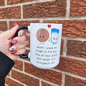 May include: White ceramic mug with a black handle. The mug features a cartoon cookie and glass of milk holding hands with a red heart above them. The text on the mug reads: "When I dunk my cookies in my milk, I think of you (& hold them under until the bubbles stop)."