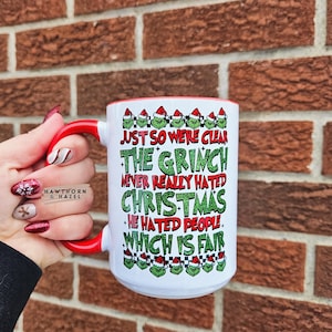 May include: White ceramic mug with a red handle and rim, featuring the text "JUST SO WERE CLEAR THE GRINCH NEVER REALLY HATED CHRISTMAS - HE HATED PEOPLE WHICH IS FAIR" in green and red. The mug has a Grinch-themed design.