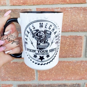 May include: White ceramic mug with a black rim and handle. The mug features a black and white graphic of a motorcycle engine with the text "Badass Mechanic Fixin' Your Shit Since Forever".