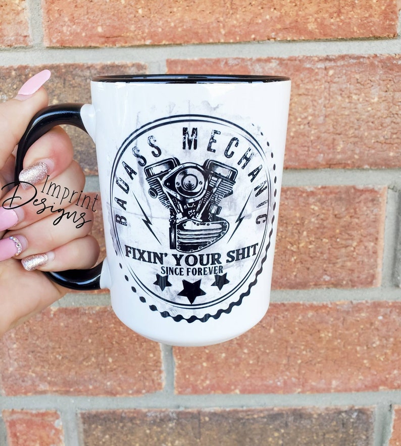 May include: White ceramic mug with a black rim and handle. The mug features a black and white graphic of a motorcycle engine with the text "Badass Mechanic Fixin' Your Shit Since Forever".