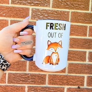 May include: White ceramic mug with a black rim and handle. The mug features a cartoon fox graphic and the text "FRESH OUT OF".