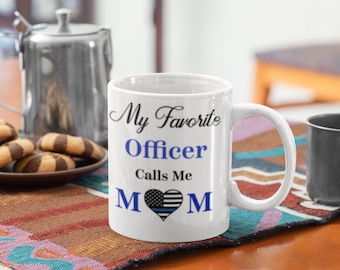 May include: White ceramic coffee mug with a blue and black thin line American flag heart design. The text on the mug reads "My Favorite Officer Calls Me MOM".