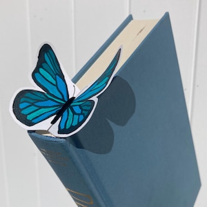 May include: A blue hardcover book with a butterfly bookmark. The butterfly has blue and teal wings with black outlines. The book is a muted blue color, and the bookmark is placed between the pages.