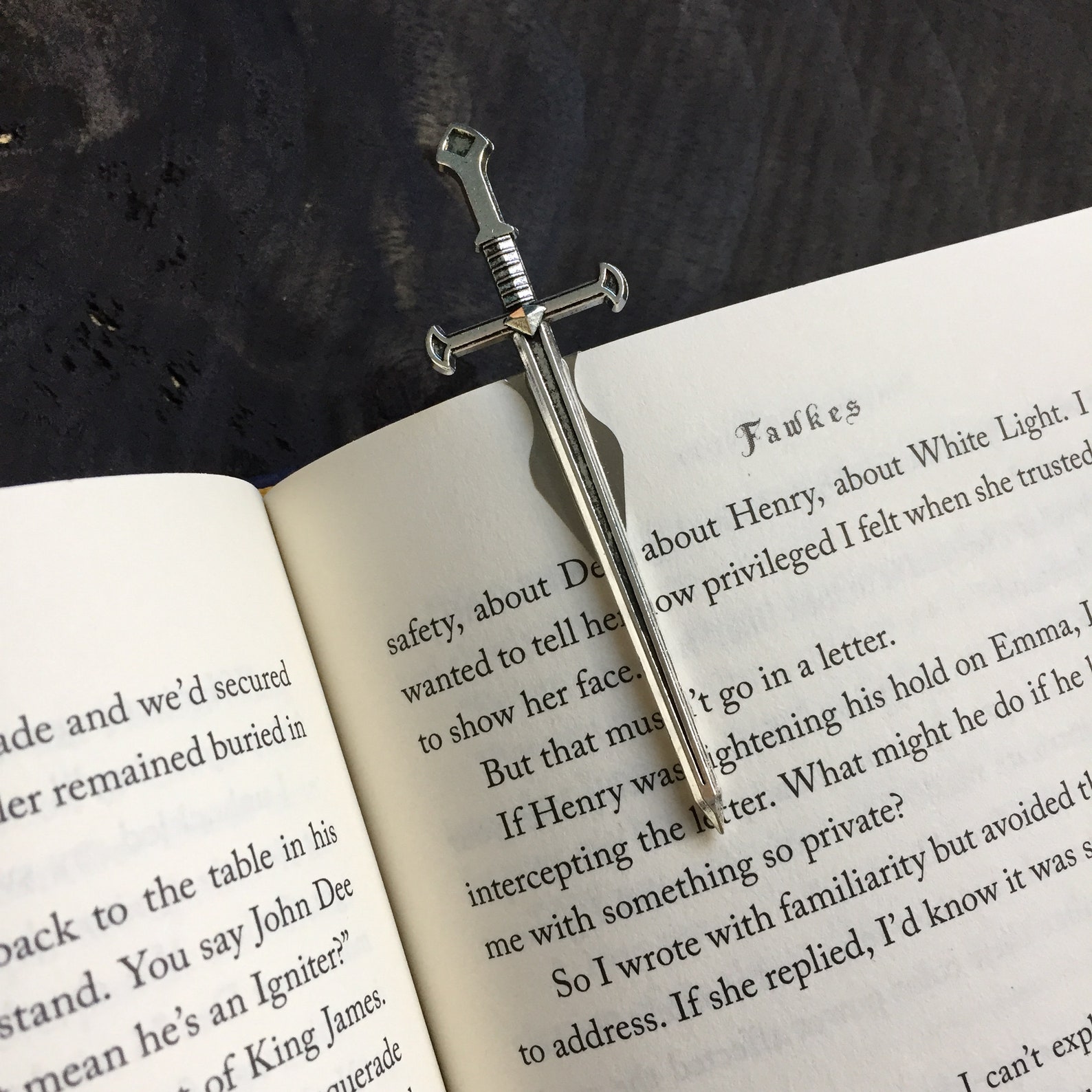 A metal sword hilt sticks out from the top of the page of a book. The front of the blade of the sword runs along the page as a bookmark.