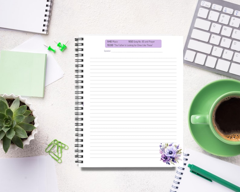 Op de afbeelding: Een spiraalgebonden notitieboek met gelinieerde pagina's, een paars-wit bloemmotief en een schema bovenaan afgedrukt. Een kopje koffie, een toetsenbord en een vetplant zijn ook zichtbaar.