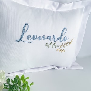 Puede incluir: Almohada blanca con el nombre "Leonardo" bordado en hilo azul. Un pequeño diseño floral bordado en verde y dorado acentúa el nombre. La almohada tiene una apariencia limpia.