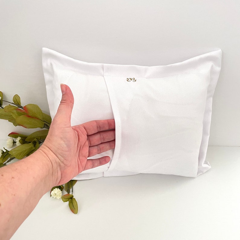 May include: White pillow with a zipper closure on the back. The zipper is visible and open, showing the inside of the pillow. The pillow has a small embroidered design on the back.