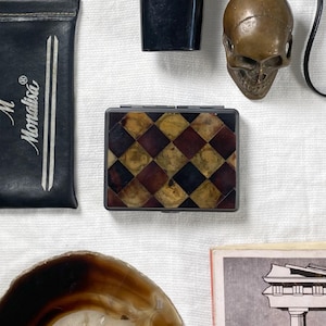 May include: An assortment of vintage items including a black and silver Super 8 film camera, a small bronze skull, a black leather case with "Monalisa" text, a checkered box, a geode slice, and a book titled "Lessons in Stoicism".