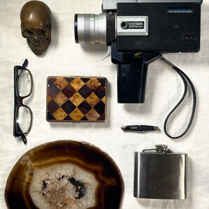 May include: An assortment of vintage items including a film camera, a skull, eyeglasses, a cigarette case, a pocket knife, a flask, a geode slice, and a gemstone. The text "TROPICAL JAVAL" and "SINATRA ROSE CIGARETTE CASE" are visible.