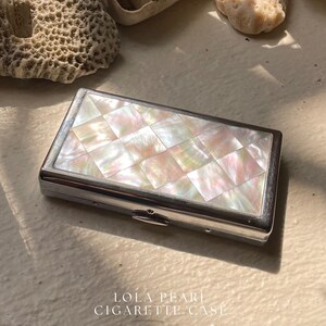 Può includere: Un portacigarette rettangolare con coperchio intarsiato in madreperla. La custodia ha una cornice in metallo color argento e una chiusura. Il testo "LOLA PEARL CIGARETTE CASE" è stampato sulla superficie.
