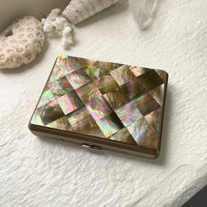 May include: A rectangular compact with a shimmering, iridescent mosaic top. The mosaic is made of small, square tiles in shades of gold, pink, and green. The compact has a gold-coloured metal frame and a small clasp. Decorative items are in the background.