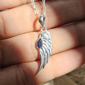 K&ouml;nnte beinhalten: Silberfarbene Halskette mit einem Anh&auml;nger in Fl&uuml;gelform und einem blauen Edelstein. Das Fl&uuml;geldesign ist detailliert. Die Halskette h&auml;ngt an einer feinen Silberkette, ein stilvolles Schmuckst&uuml;ck.