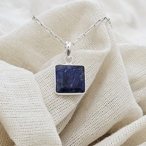 Puede incluir: Un collar de plata con un colgante cuadrado de color azul oscuro. El colgante está engastado en un marco de plata y cuelga de una fina cadena de plata. El collar descansa sobre una tela crema texturizada.