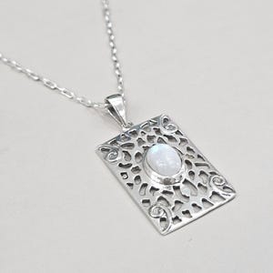 Può includere: Una collana con pendente in argento con un pendente rettangolare con una gemma ovale e iridescente. Il pendente ha un design traforato ed è sospeso a una delicata catena. La collana è mostrata su una superficie grigio chiaro.