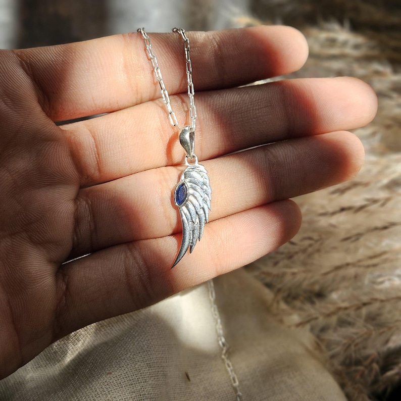 K&ouml;nnte beinhalten: Silberfarbene Halskette mit einem Anh&auml;nger in Fl&uuml;gelform und einem blauen Edelstein. Das Fl&uuml;geldesign ist detailliert. Die Halskette h&auml;ngt an einer feinen Silberkette, ein stilvolles Schmuckst&uuml;ck.