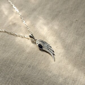 K&ouml;nnte beinhalten: Silberfarbene Halskette mit einem Anh&auml;nger in Fl&uuml;gelform und einem dunkelblauen Edelstein. Der Fl&uuml;gelanh&auml;nger h&auml;ngt an einer feinen Kette. Ein elegantes Schmuckst&uuml;ck mit symboltr&auml;chtigem Design.