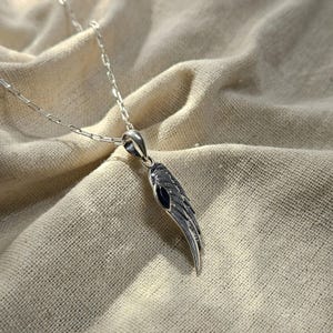 K&ouml;nnte beinhalten: Halskette mit silbernem Fl&uuml;gelanh&auml;nger. Der Anh&auml;nger hat ein detailliertes Fl&uuml;geldesign mit dunkler, strukturierter Oberfl&auml;che und aufwendigen Federdetails. Ein kleiner, dunkelblauer Akzent ist sichtbar. Die Silberkette liegt auf beigem Stoff.