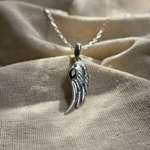 K&ouml;nnte beinhalten: Eine silberfarbene Halskette mit einem Fl&uuml;gelanh&auml;nger und einem dunkelblauen Edelstein. Der Fl&uuml;gelanh&auml;nger ist mit federartigen Linien versehen und h&auml;ngt an einer filigranen Kette. Die Halskette wird auf einem strukturierten, neutralfarbenen Stoff pr&auml;sentiert.