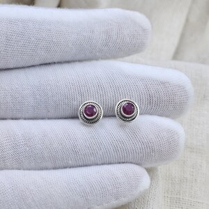 May include: A pair of round stud earrings featuring a central circular dark pink gemstone set in a silver bezel. The silver setting is decorated with a twisted rope-style border around the edge of each earring, creating a textured metallic frame.