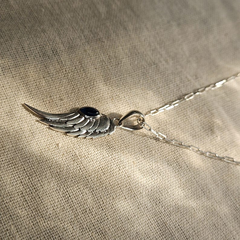 K&ouml;nnte beinhalten: Halskette mit silberfarbenem Anh&auml;nger in Form eines detaillierten Fl&uuml;gels mit einem kleinen, dunkelblauen Stein. Der Fl&uuml;gelanh&auml;nger ist an einer filigranen Kette befestigt.