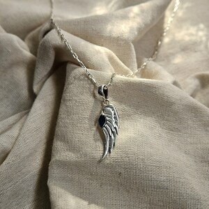 K&ouml;nnte beinhalten: Eine silberfarbene Halskette mit einem Anh&auml;nger in Form eines Fl&uuml;gels. Der Fl&uuml;gel hat detaillierte Federstrukturen und einen kleinen, dunkelblauen Stein. Die Kette ist filigran und silberfarben, auf einem neutralfarbenen Stoff.