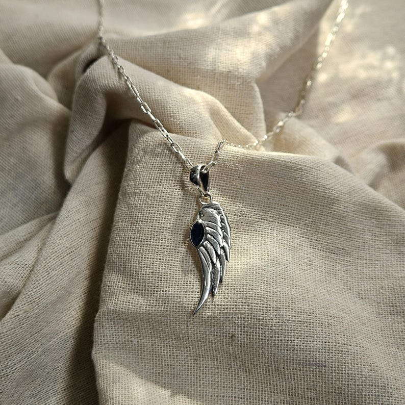 K&ouml;nnte beinhalten: Eine silberfarbene Halskette mit einem Anh&auml;nger in Form eines Fl&uuml;gels. Der Fl&uuml;gel hat detaillierte Federstrukturen und einen kleinen, dunkelblauen Stein. Die Kette ist filigran und silberfarben, auf einem neutralfarbenen Stoff.